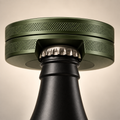 Close-up of a bottle opener snus tin with a matte black bottle and an olive green colour, shown partially inserted into a bottle neck against a warm beige background.