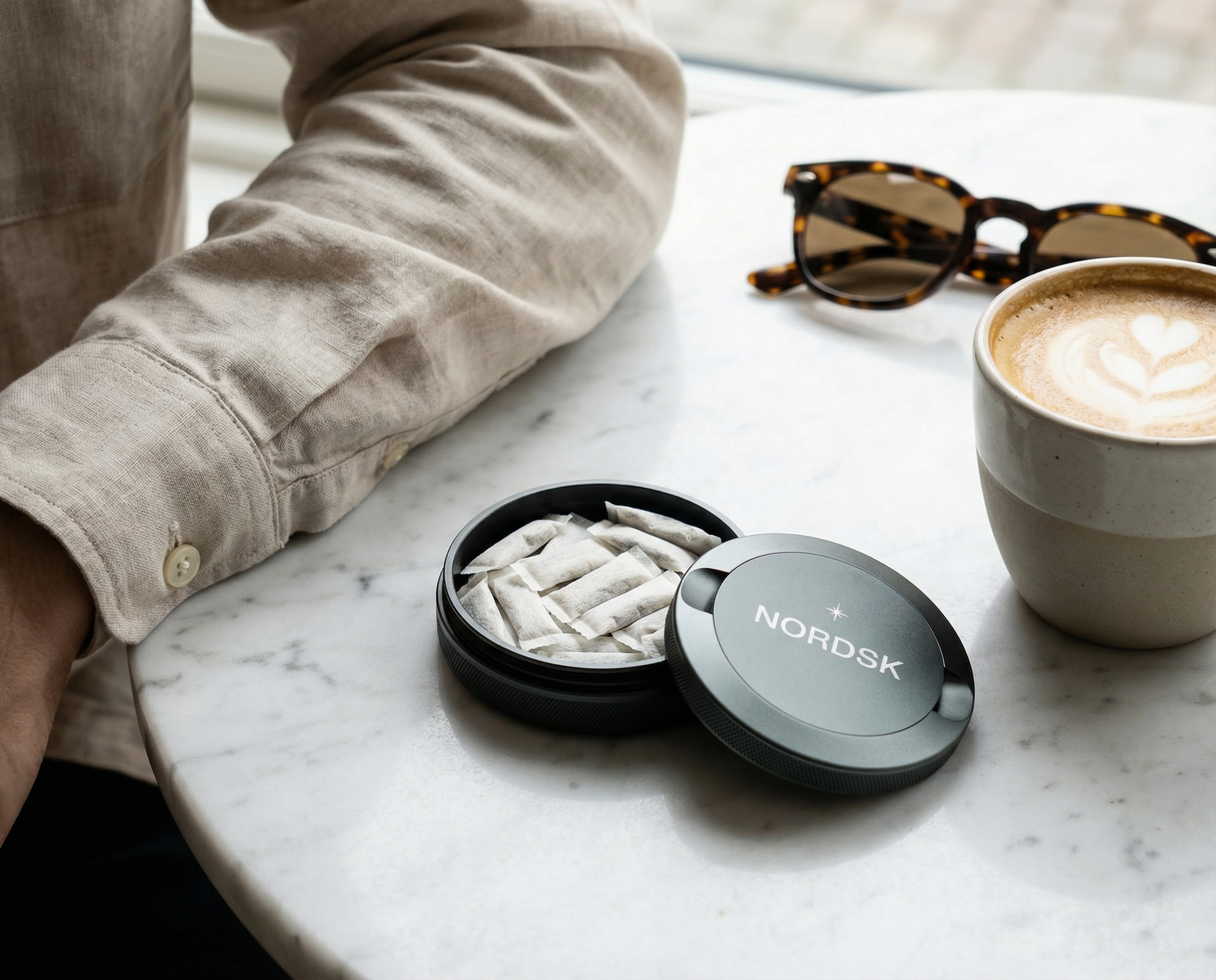 A person in a beige linen shirt sits at a white marble table next to a cup of latte art coffee, tortoiseshell sunglasses, and an open round black snusdosa container from NORDSK with snus inside.