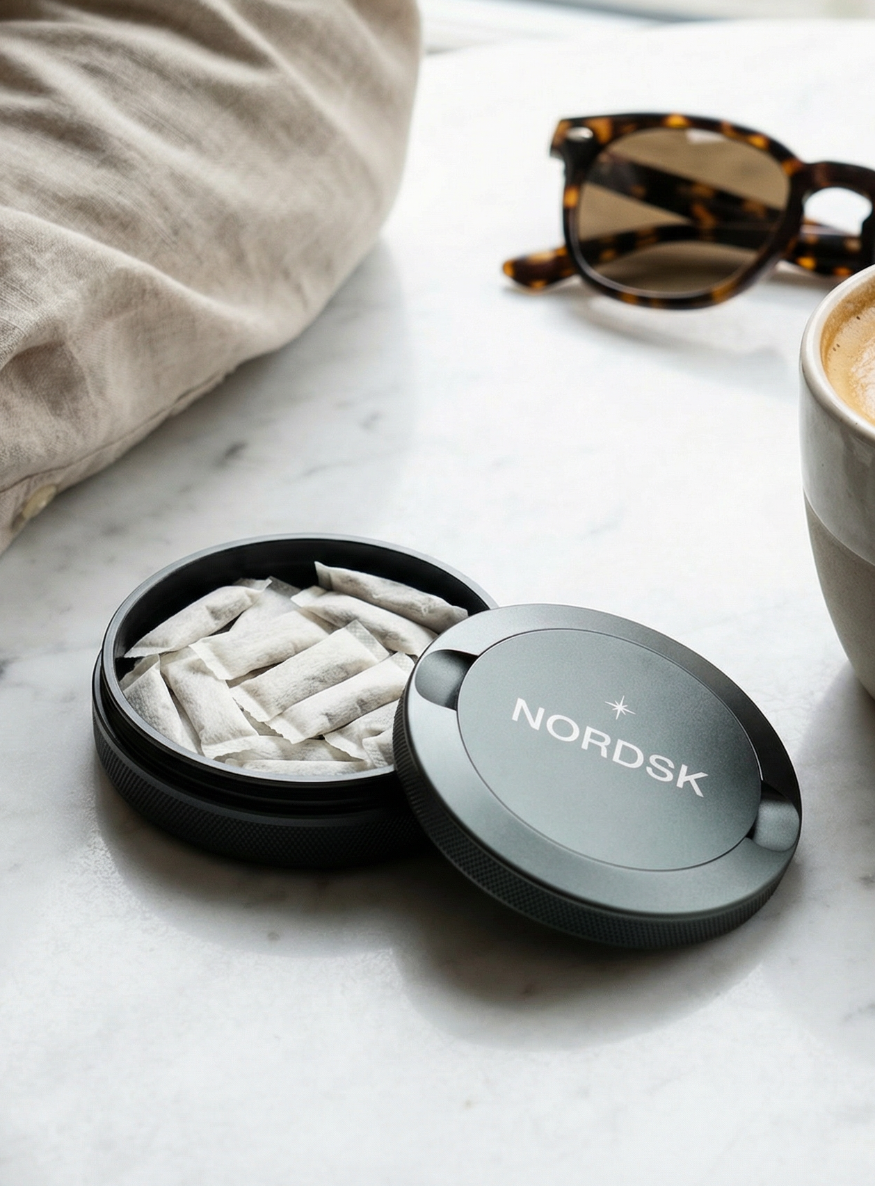 A person in a beige linen shirt sits at a white marble table next to a cup of latte art coffee, tortoiseshell sunglasses, and an open round grey snus container from NORDSK.